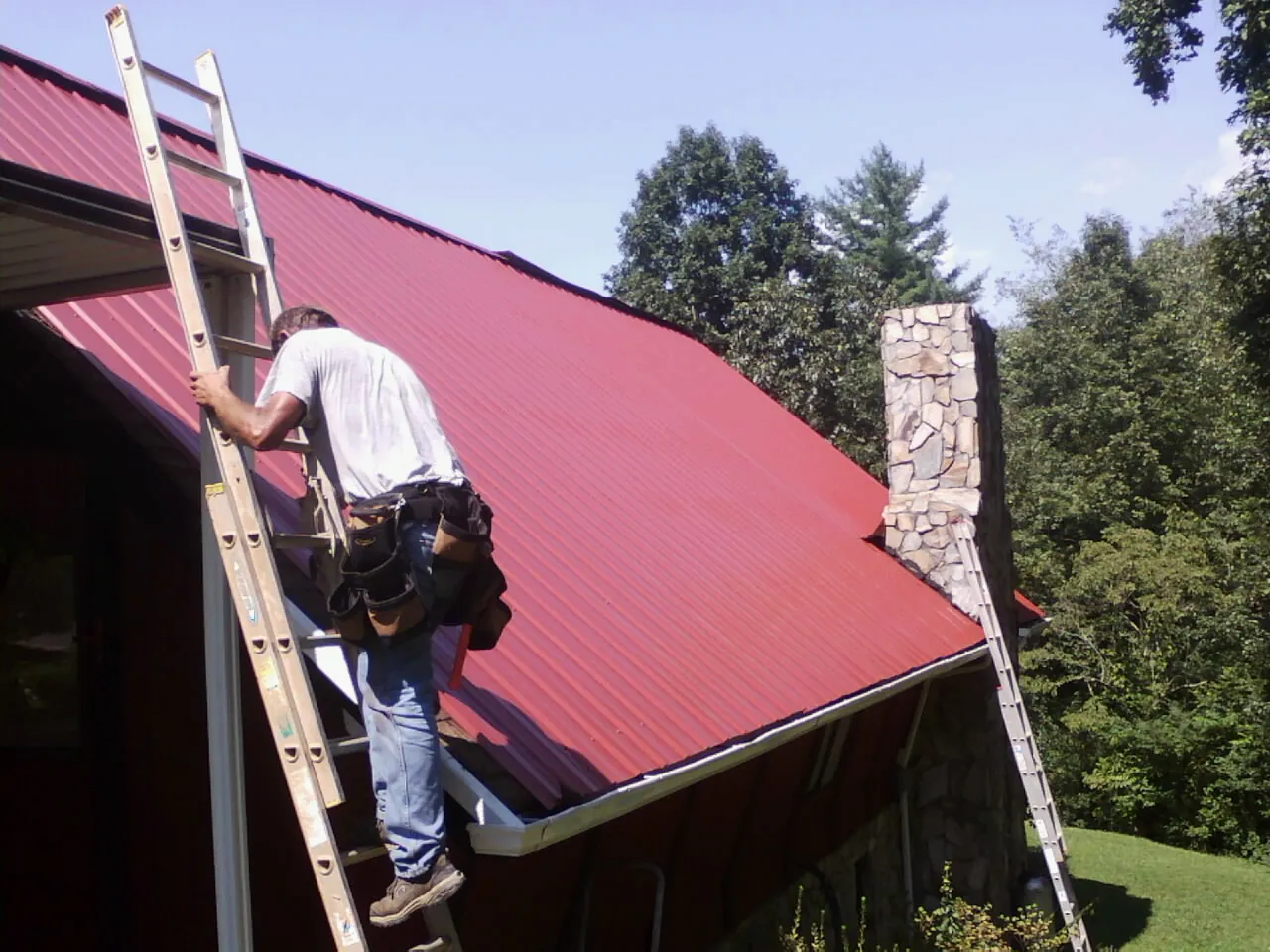 Roofer on a ladder beside a red metal roof and stone chimney — Soffit & Fascia Repair by Roofing Brothers in Greenview